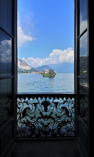 Immagine con lago, nuvole, cielo, montagne, isola, acqua, finestra, ringhiera, balcone, ferro, azzurro, vetri, porta, affaccio, cornice, porte, case, riflesso, riflessi, panorama, mare, inferriata, vista, isolotto