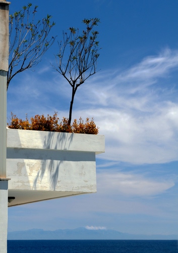 Immagine con nuvole, cielo, bianco, piante, mare, balcone, azzurro, montagne, blu, palazzo, ombra, fiori, alberi
