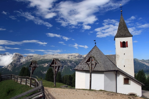 Immagine con chiesa, nuvole, campanile, croci, cielo, staccionata, montagne, montagna, azzurro, legno, sentiero, verde, bianco, strada, erba, crocifissi