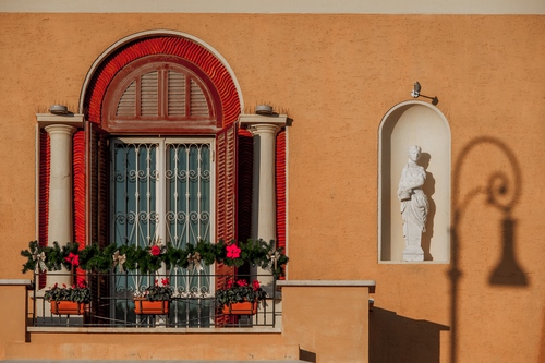 Immagine con statua, fiori, balcone, ombra, finestra, colonne, rosso, vasi