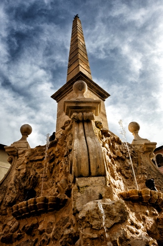 Immagine con nuvole, cielo, fontana, acqua, obelisco, zampilli, sfere, azzurro, marrone, prospettiva