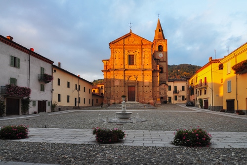 Immagine con chiesa, piazza, fontana, case, campanile, fiori, cielo, vasi, nuvole, finestre, balconi, orologio, porte, fioriere, piante