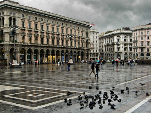 Immagine con piazza, piccioni, pioggia, ombrelli, palazzi, nuvole, persone, portici, archi, finestre, lampioni, colonne, cielo, bagnato, città, colombi