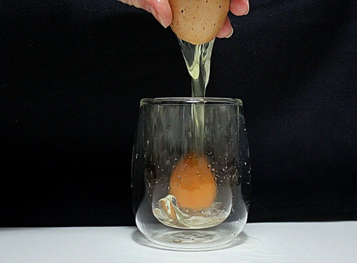 Immagine con bicchiere, uovo, tuorlo, albume, guscio, mano, nero, dita, bianco, vetro, crudo, trasparenza, giallo, rotto, tavolo