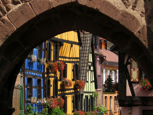 Immagine con arco, giallo, finestre, fiori, azzurro, colori, case, vasi, mattoni, piante, rosso, tetti, pietre, bianco, marrone, tegole, blu, fioriere